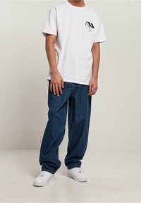 Camiseta blanca de algodón con un pequeño gráfico negro en el pecho, combinada con unos vaqueros azules sueltos y zapatillas blancas.