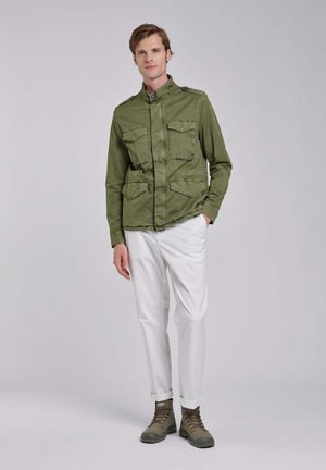 Jonge man staand, gekleed in een groene militair-stijl jas, witte opgerolde broek en bruine veterlaarzen, tegen een effen achtergrond.