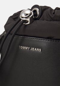 Czarna torba z gładką skórzaną podstawą i marszczonym materiałowym wierzchem, zabezpieczona metalowym sznurkiem ściągającym i srebrnymi elementami. Wytłoczone logo "TOMMY JEANS".