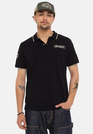 Homme portant un polo noir Von Dutch, une casquette assortie Von Dutch, un jean foncé, avec des tatouages de papillon et d’araignée sur ses avant-bras.