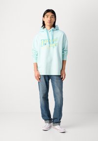 Lyseblå hoodie med "Level Up" grafik i gul og blå. Sammen med slidte blå jeans og hvide sneakers. Blød, afslappet design.
