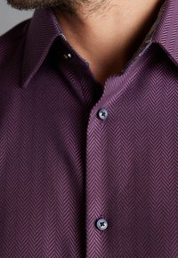 Nahaufnahme eines lila Fischgrätmusters auf einem Hemd mit schwarzen Knöpfen, getragen von einer Person mit heller Haut und Gesichtsbehaarung.