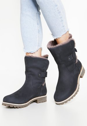 Personne portant des bottes d'hiver noires mi-mollet avec doublure en fourrure et boucles, associées à un jean court bleu clair avec des ourlets effilochés.