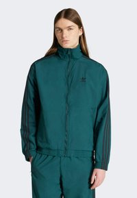 Teal træningsjakke med høj krave, lynlåslukning og sorte tre-striber. Stoffet ser glat ud med en elastisk talje.