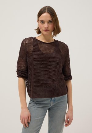 Jonge vrouw met kort haar, gekleed in een bruin opengebreid vest over een zwarte top, gecombineerd met lichtblauwe spijkerbroek, staand tegen een effen achtergrond.