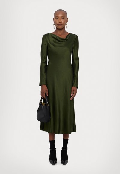 Groene satijnen jurk met een gedrapeerde halslijn, lange mouwen en een uitlopende zoom, gecombineerd met een kleine zwarte tas en zwarte puntige hakken.
