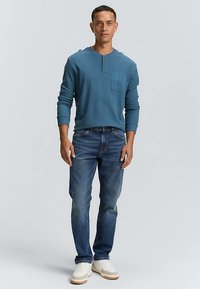 Sinine pikkade varrukatega henley särk ees taskuga, kombineeritud tumedate teksapükste ja valgete tossudega. Sile tekstuur ja kehakuju rõhutav lõige.