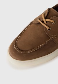 Bruine suede bootshoe met tan leren veters, zichtbare witte stiksels en een doorschijnende off-white rubberen zool.