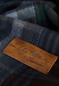 Wollen stof in een donkergroen en blauw ruitpatroon met een bruine leren label waarop handgeschreven tekst en geperste details staan.