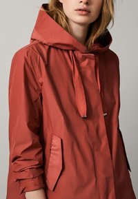 Vattenavvisande röd jacka med huva, med dragkedja, dragsko och två sidofickor. Slät textur och modern design.