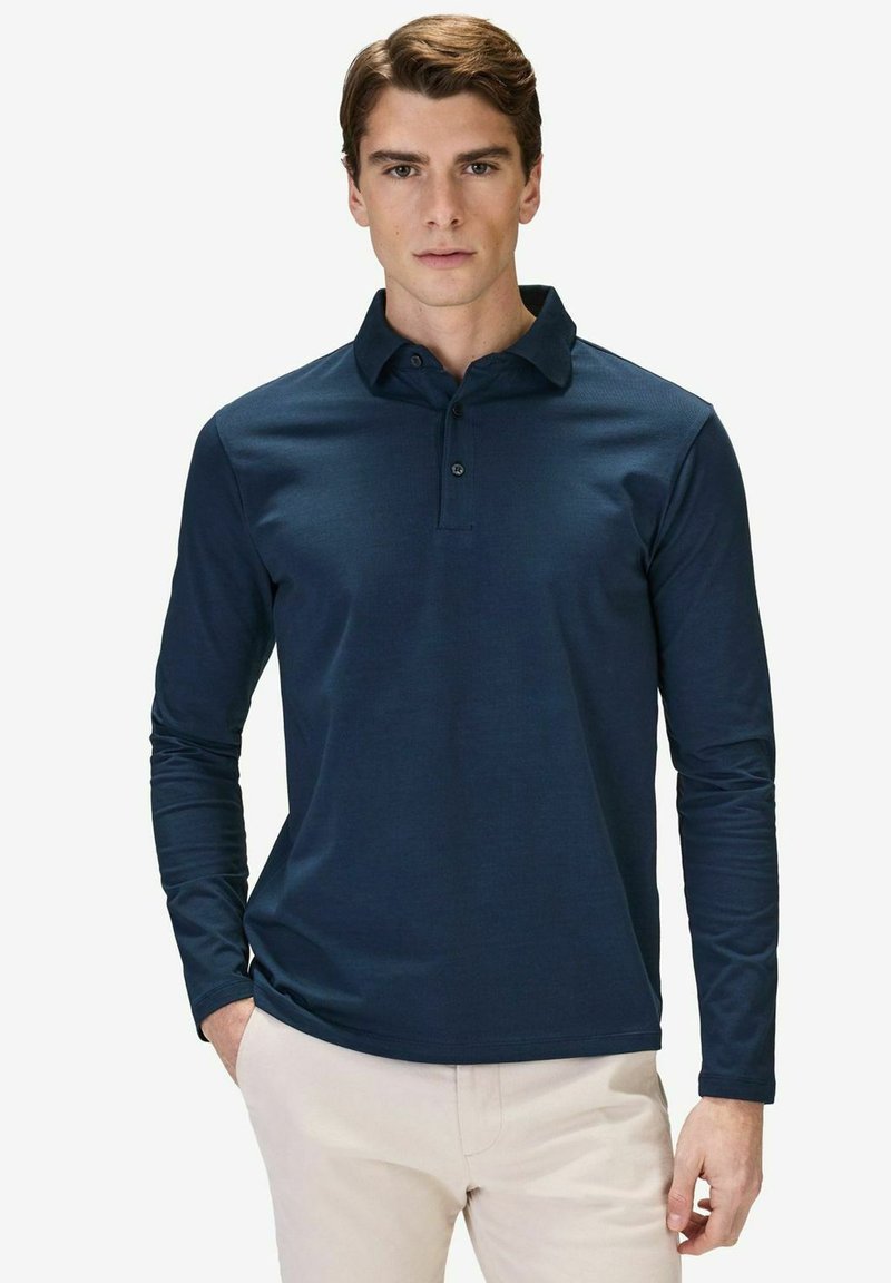 Mörkblå långärmad polo med krage, två knappar i placket, slät textur, kombinerad med ljusa byxor, händernas i fickorna.