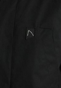 Juodas švarkas su tekstūruotu audiniu, turintis mažą pleistrą, rodančią baltą abstraktų dizainą. Dizainas yra krūtinės kišenės srityje.