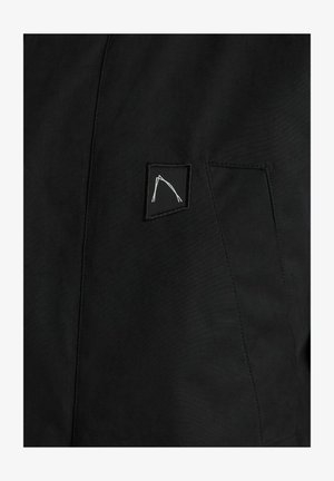 Černá bunda s texturovanou tkaninou, vybavená malou nášivkou s bílým abstraktním vzorem. Vzor se nachází v oblasti kapsy na prsou.