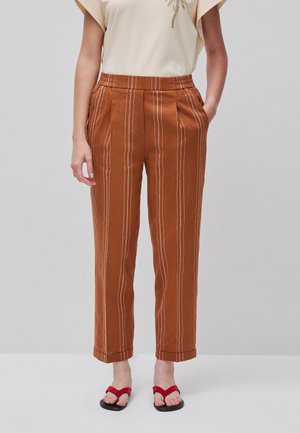 Femme portant un pantalon taille haute couleur rouille avec des rayures verticales blanches et des tongs rouges, debout les mains dans les poches.