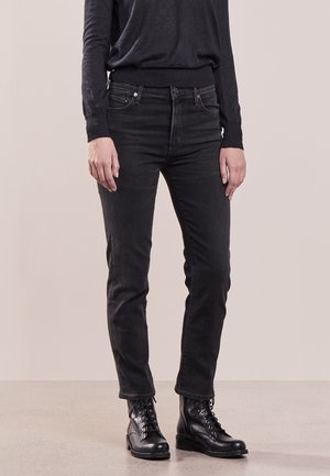 Person, der står iført sorte jeans, sort langærmet top og sorte snøreløse ankelstøvler på en lys gulv og neutral vægbaggrund.