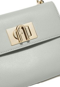 Lichtgrijze leren handtas met een opvallende gouden metalen druksluiting en fijne textuur, met gedetailleerde stiksels langs de randen.