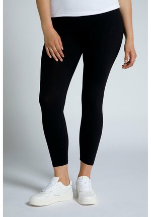 Schwarze Leggings aus dehnbarem Stoff, kombiniert mit weißen Sneakers. Das Design zeichnet sich durch eine enganliegende Silhouette und eine glatte Textur aus.