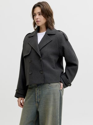 Noor naine, kes kannab tumedat halli ruudulist topeltsõlmusega lühikest jopet valge särgi ja lahtiste tuhmistunud siniste teksade üle, seisab ühe käega taskus.