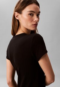 Woman with straight brown hair wearing a black short-sleeve shirt, turned slightly away, looking back over her shoulder against a plain gray background.