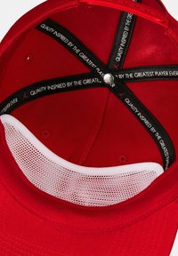 De binnenkant van de rode baseballcap heeft een witte meshvoering en een zwarte verstelbare band met de tekst "KWALITEIT GEÄNSPIREERD DOOR DE GROOTSTE SPELER OOIT."