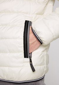La giacca puffer bianca presenta un design trapuntato, tasche con zip nere con dettagli a righe e polsini a coste. Texture liscia e lucida.