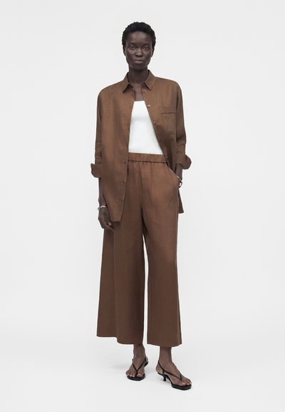 Γυναίκα στέκεται φορώντας καφέ παντελόνια με φαρδύ πόδι και αντίστοιχο καφέ πουκάμισο με κουμπιά πάνω από λευκή φανέλα και μαύρες ψηλοτάκουνες με λουράκια.