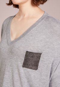 Grau gestrickter Pullover mit V-Ausschnitt, der eine strukturierte schwarze Tasche auf der linken Seite aufweist. Weiches Material mit lässigem Schnitt und kurzen Ärmeln.