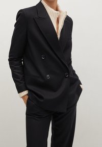 Personne portant un blazer noir croisé sur un pull côtelé crème, les mains dans les poches, sur un fond clair uni.