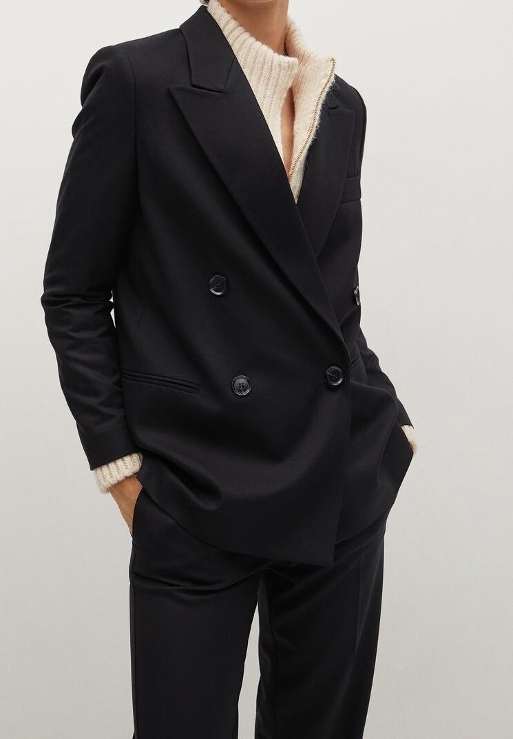 Personne portant un blazer noir croisé sur un pull côtelé crème, les mains dans les poches, sur un fond clair uni.