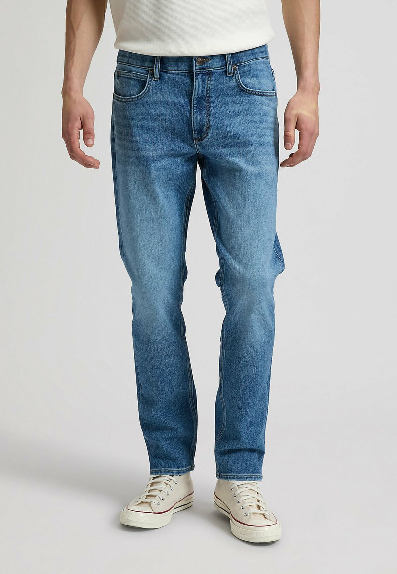 Homme portant un jean slim bleu clair et des baskets basses blanches, debout devant un fond uni.