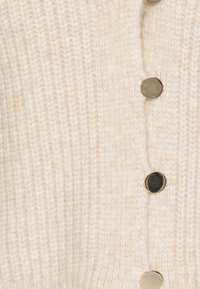 Stickad kofta i off-white med ett texturerat mönster och framträdande knappar i guldton längs framsidan för stängning.