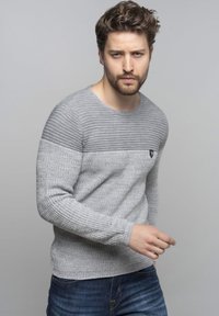 Pull en maille grise avec texture côtelée, présentant un motif de rayures horizontales sur les épaules et un petit logo sur la poitrine.