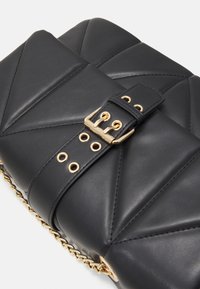 Sac à main en cuir matelassé noir avec boucle dorée et œillets sur la sangle, et détails en chaîne dorée sur fond blanc.