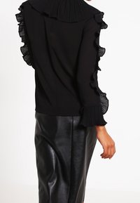Blusa nera con colletto plissettato e bordi delle maniche arricciati, abbinata a una gonna in pelle nera aderente. Texture liscia in tutto.