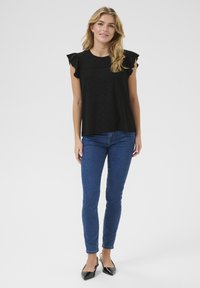 Haut noir à manches à volants avec un motif texturé, associé à un jean skinny bleu et des escarpins noirs à bout pointu. Design simple et décontracté.