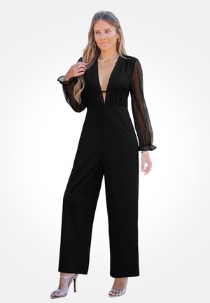 Kobieta w czarnym kombinezonie z przezroczystymi, długimi rękawami, głębokim dekoltem w kształcie litery V, stojąca z jedną ręką dotykającą włosów i srebrnymi sandałami na wysokim obcasie.