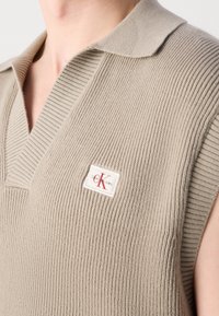 Beige gestricktes ärmelloses Top mit V-Ausschnitt und geripptem Muster. Verfügt über ein Calvin Klein Logo-Patch auf der linken Seite.