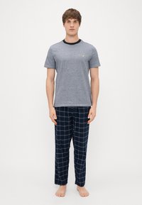 T-shirt gris à manches courtes avec un col noir et un petit emblème, associé à un pantalon de pyjama à carreaux bleu foncé. Pas de chaussures.
