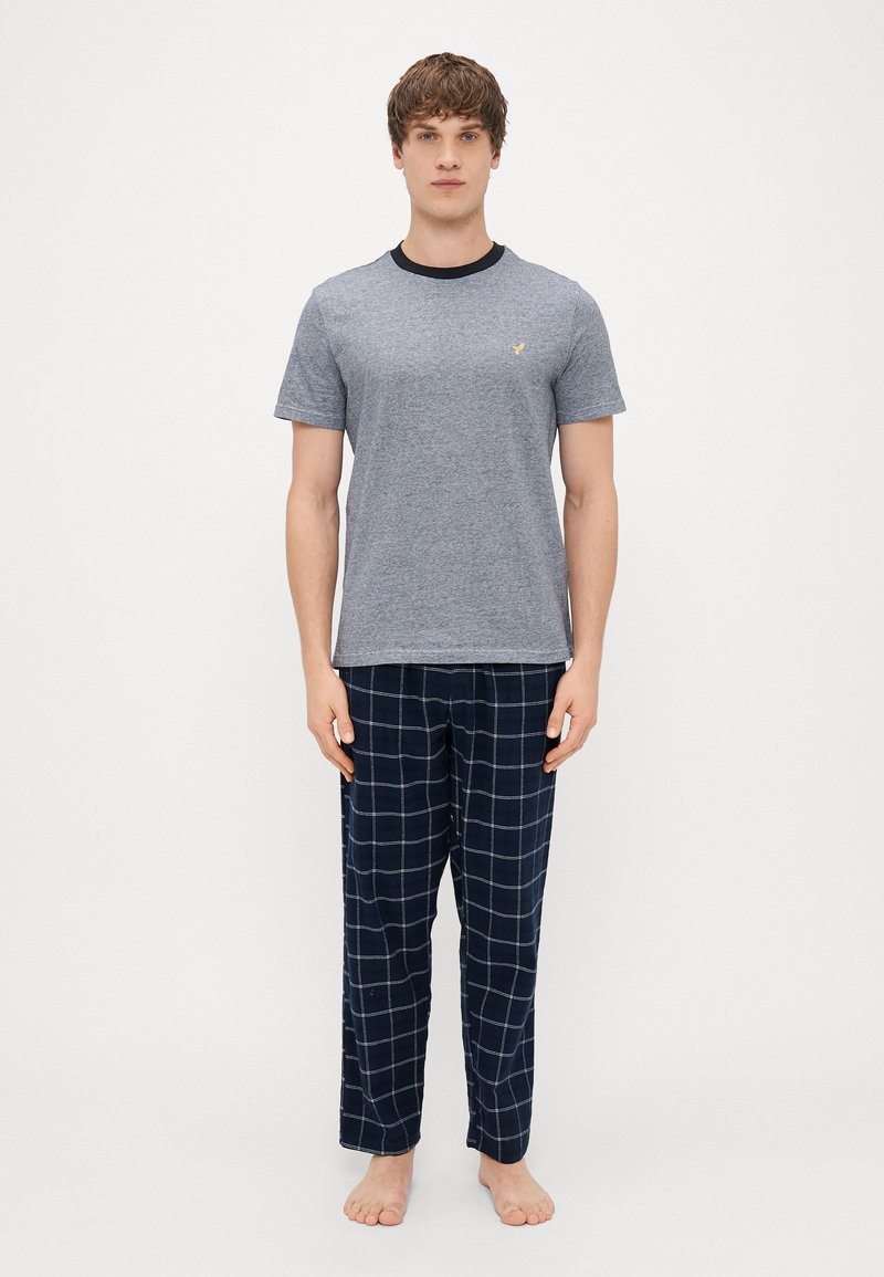 T-shirt gris à manches courtes avec un col noir et un petit emblème, associé à un pantalon de pyjama à carreaux bleu foncé. Pas de chaussures.