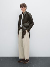 Dunkelgrüne Lederjacke mit Reißverschluss und Seitentasche, beigefarbene weite Hose, braune Wildlederschuhe und Sonnenbrille. Glatte Textur.
