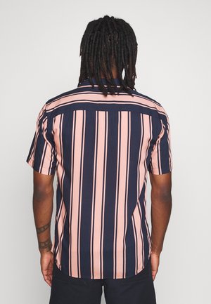 Homme avec dreadlocks portant une chemise à manches courtes rayée verticalement bleu marine et rose, tourné de dos sur un fond uni.
