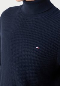 Gros plan d'une personne portant un pull à col roulé en maille texturée bleu marine avec un petit logo brodé rouge et blanc sur la poitrine.