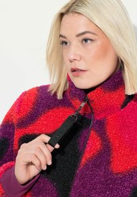 Veste en polaire avec des motifs rouges, violets et noirs ; col montant, fermeture éclair avec tirette, tissu texturé avec une apparence épaisse et moelleuse.