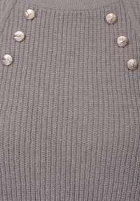 Strikket genser i lys grå med ribbestruktur. Har fire dekorative gullknapper på skulderen som gir et subtilt preg.