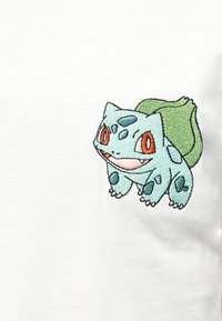 Μικρό κεντημένο πλάσμα σε μπλε-πράσινο χρώμα με μεγάλα πορτοκαλί μάτια, κηλίδες και ένα πράσινο μπουμπούκι στην πλάτη του σε λευκό ύφασμα.