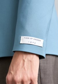 Svijetloplava tkanina rukava s bijelom etiketom na kojoj piše "TIGER OF SWEDEN 1903." Ističe se glatkom teksturom i prilagođenim krojem.