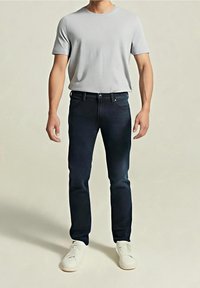 Homme portant une chemise à manches courtes gris clair, un jean bleu foncé et des baskets blanches, debout devant un fond clair uni.