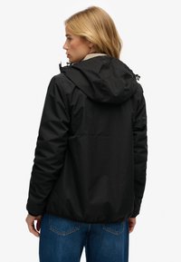 Sort vandtæt jakke med hætte, med et enkelt design, elastiske manchetter og en glat tekstur. Bruges over blå jeans.