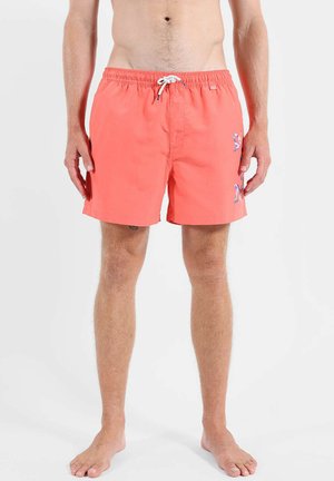 Homme portant un short de bain corail avec un cordon blanc et un petit logo à motif sur la jambe droite, debout pieds nus sur un fond blanc.