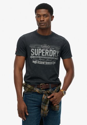 Black cotton T-shirt with a white graphic print featuring "SUPERDRY" text. Worn with blue jeans and a green plaid shirt wrapped around the waist.